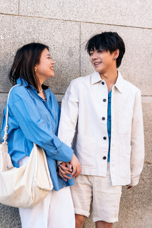 Young asian couple standing against gray wall laughing and looking at each other.