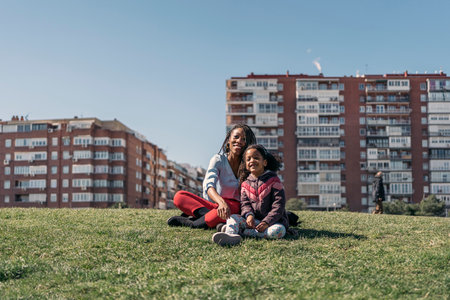 Cute afro girl sitting in the grass with her mom, smiling, looking at camera and enjoying sunny day.の写真素材