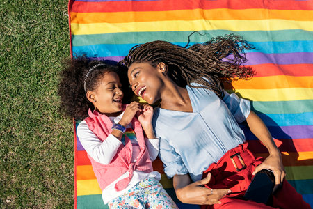 Cute afro girl lying down in the grass in colorful picnic blanket with her mom and enjoying sunny day.の写真素材