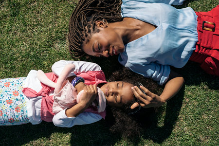 Adorable afro girl hugging stuffed animal lying down in the grass with her mother. They are smiling and having fun.の写真素材