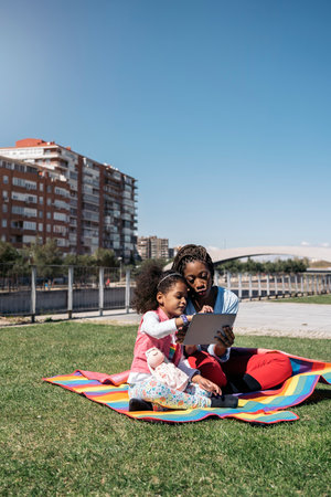 African woman sitting in the grass with her young daughter and using tablet. They are enjoying a sunny day.の写真素材