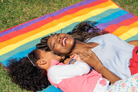 Cute afro girl lying down in the grass in colorful picnic blanket with her mom and enjoying sunny day.の写真素材