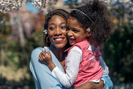 Black woman with braids hugging her young daughter in the park during spring sunny day.の写真素材