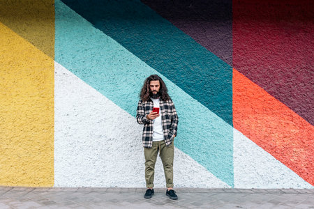 Confident hipster man standing against colorful wall in the city and using his phone.の写真素材