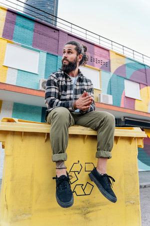Cool hipster man with long beard sitting in a recycling container and looking to the side.