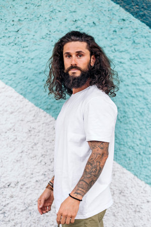 Attractive bearded man with long hair posing and looking at camera against blue wall.