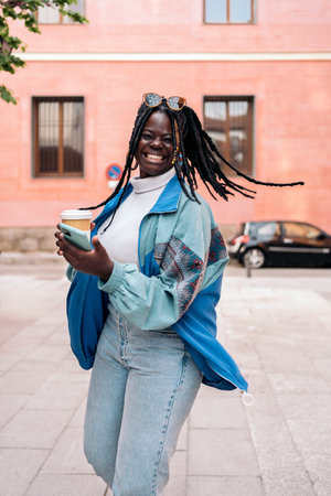 Cheerful black girl with cool braids having fun in the city. She is holding her mobile phone and a coffee cup.の写真素材