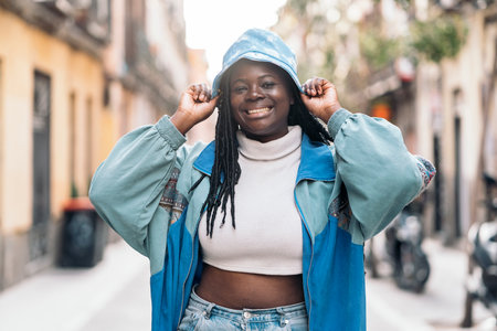 Cool young african woman with braids smiling and walking in the city. She is wearing casual clothes and a hat.の写真素材