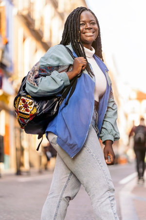 Happy and young african woman with cool braids smiling and walking in the city. She is using a...
