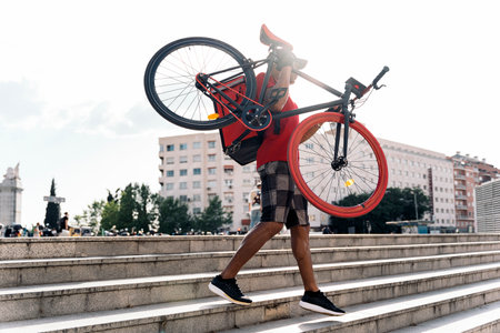 Unrecognized messenger man wearing cap carrying his bike in the city on the way for delivering a package.の写真素材