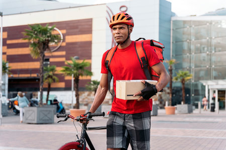 African delivery man wearing safety helmet carrying his bike in the city on the way for delivering a package.の写真素材