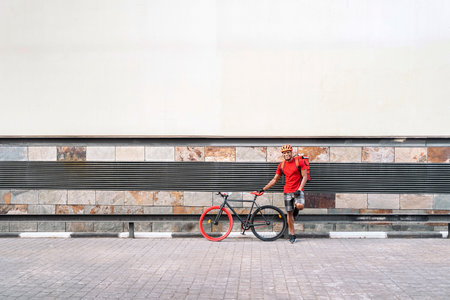 African american delivery man wearing safety helmet smiling next to his bike and looking at camera in the street.の写真素材