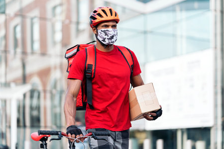 African delivery man wearing face mask carrying his bike in the city on the way for delivering a package.の写真素材