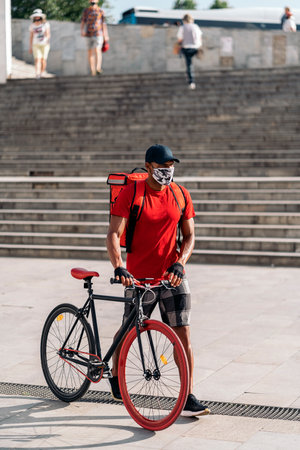 African delivery man wearing face mask carrying his bike in the city on the way for delivering a package.の写真素材