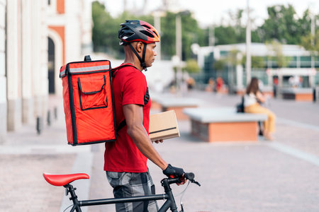 African delivery man wearing safety helmet standing with his bike in the city and holding a package.の写真素材