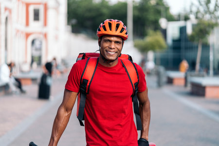 African american delivery man wearing safety helmet smiling and looking at camera in the street.の写真素材