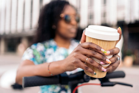 Unrecognized african woman with her nails done holding a coffee cup while standing in her electric scooter.の写真素材