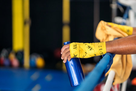Relaxed young boxer leaning on the ropes of the ring drinking water. Concept of training.
