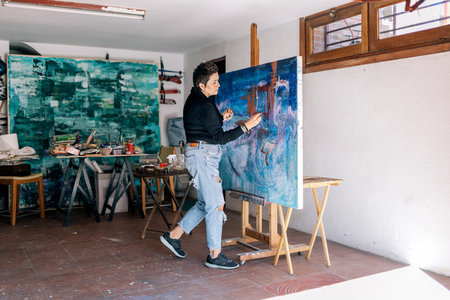 Middle aged artist working in her garage painting on a big canvas with blue paint. She is wearing...