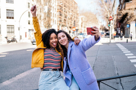 Beautiful young girls with curly hair smiling and taking selfie with mobile phone in the street.の写真素材