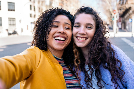 Beautiful young girls with curly hair smiling and taking selfie on the street.