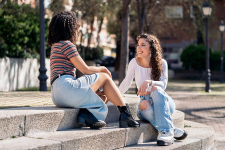 Curly haired girls sitting in the street looking at each other and talking. They are smiling and having fun.の写真素材