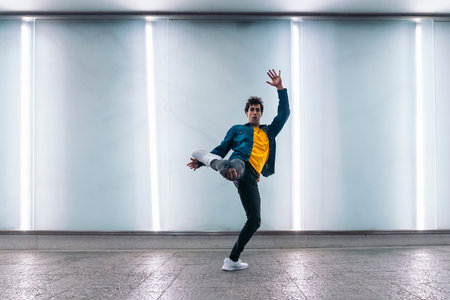 Talented young man practicing break dance against white wall with lights and looking at camera.
