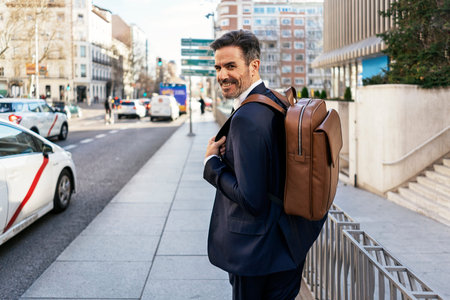 Back view of mature male entrepreneur with leather backpack walking along street and commuting to...
