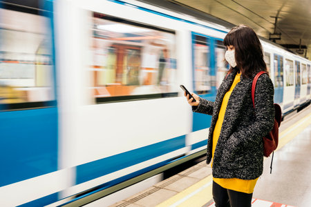 Stock photo of adult woman wearing face mask due to covid19 using her phone while the metro arrives.の写真素材