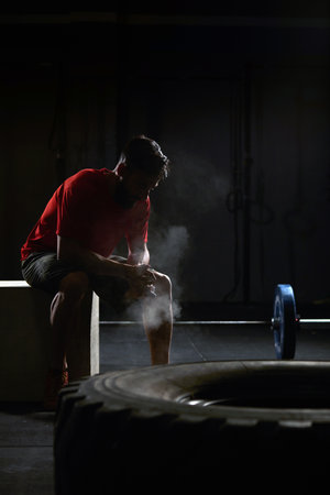 Stock photo of an adult man in a gym sitting down and resting. There is a tractor tire in front and a barbell behind him. He is wearing sportswear.の写真素材
