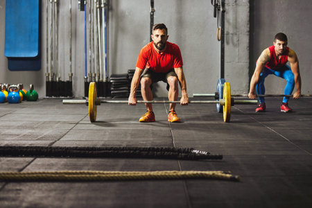 Stock photo of two adult men in a gym squatting down and lifting barbells. They are wearing...