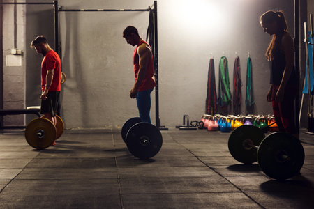 Stock photo of three adults in a gym squatting down and lifting barbells. They are wearing sportswear. There are dumbbells in the background.の写真素材