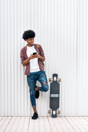 Stock photo of cool african boy standing next to his longboard smiling and using cellphone.の写真素材