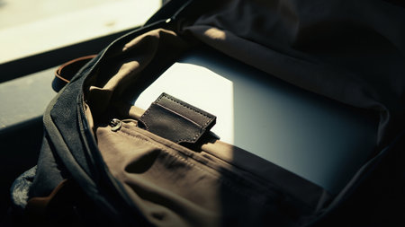 A close-up, sunlit view showcases an open backpack, revealing a sleek laptop and a sophisticated leather wallet. The image highlights essential accessories for business and travel, ideal for a modern professional's everyday carry (EDC). This detailed shot in a softly lit workspace setting emphasizes the stylish and elegant arrangement. Perfect for content creators, the image is suitable for articles on technology, minimalist design, or professional work setups.の素材