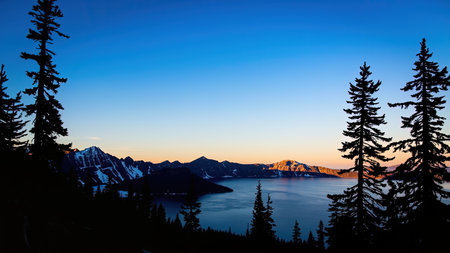 Witness the breathtaking beauty of Crater Lake, Oregon, as the sun dips below the horizon, painting the sky with vibrant hues. This stunning panoramic view showcases the lake's serene waters reflecting the silhouettes of evergreen trees and majestic mountain peaks against a clear blue sky. Ideal for travel and nature publications, this image captures the essence of wilderness adventure and tranquil exploration. The vibrant sunset creates a captivating atmosphere, making it perfect for showcasing natural beauty and peaceful landscapes.の素材