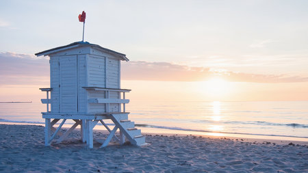 White lifeguard stand on a sandy beach at sunrise with a red flag and calm ocean water.の素材