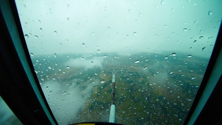 Raindrops on a cockpit window obscure the view of a green landscape under a cloudy sky.の素材