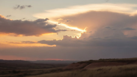 A breathtaking sunset paints the sky with vibrant hues of orange, pink, and purple. Dramatic clouds are illuminated by the setting sun, with bright rays of light breaking through. The foreground shows the soft contours of rolling hills and dry grass, creating a sense of vastness and tranquility. The scene evokes a peaceful and awe-inspiring natural spectacle.の素材