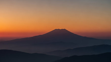 A striking silhouette of layered mountains against a vibrant sunset sky. The foreground mountains are dark and indistinct, fading into softer blue hues, while the prominent central peak stands out sharply against the warm orange and red gradient of the sky.の素材
