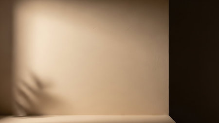 A softly lit wall with a subtle texture in a neutral beige or cream color. Diffused light casts gentle shadows, including a hint of leaf shapes, on the left side. The image features a corner where the wall meets another darker surface, creating a sense of depth. The overall impression is minimalist and clean, suitable for backgrounds or design elements.の素材