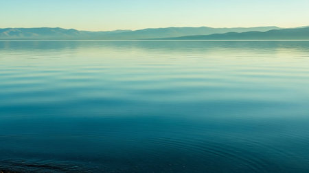 A vast and tranquil lake stretches towards a distant mountain range under a clear, pale blue sky. Gentle ripples disturb the surface of the water, creating soft patterns that reflect the serene atmosphere. The minimalist landscape evokes a sense of peace and quietude.の素材