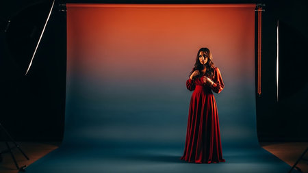 A young woman stands in a photography studio wearing a long, flowing red dress. She is positioned in front of a gradient backdrop that transitions from a warm orange at the top to a cool blue at the bottom. Studio lighting equipment is visible on either side of the frame, casting dramatic shadows and illuminating the subject. The woman is looking slightly away from the camera with her hands clasped near her chest, conveying a sense of elegance and poise.の素材