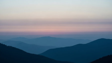 A serene landscape of layered mountain ranges fading into the distance. The mountains are depicted in deep blue silhouettes against a soft, pastel sky transitioning from pale pink to lavender and then to a light blue. A gentle mist or haze softens the distant peaks, creating an atmospheric and tranquil scene.の素材