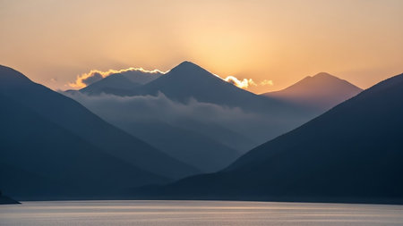Dark blue mountain peaks are silhouetted against a soft orange and yellow sunrise sky, with rays of light breaking through clouds.の素材