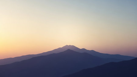 Layered mountain peaks are silhouetted against a soft gradient sky transitioning from warm yellow to cool purple at sunset.の素材