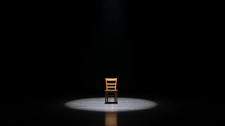 A solitary wooden chair sits in the center of a dark stage, illuminated by a single, focused spotlight, creating a stark and dramatic contrast.の素材