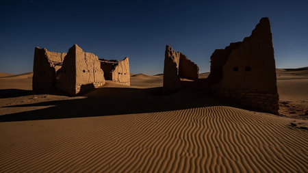 Ancient, crumbling mud brick ruins stand amidst rippling sand dunes under a clear night sky, casting long shadows and evoking a sense of history.の素材