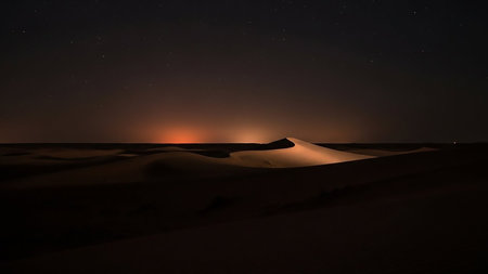 Silhouetted desert dunes are softly illuminated by a warm, distant light on the horizon under a dark, star-filled night sky.の素材