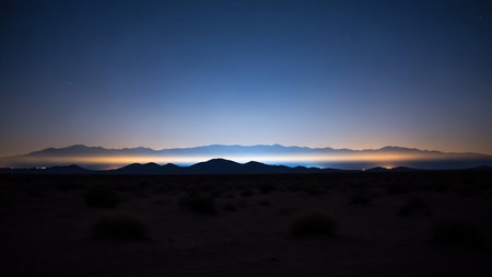 A wide desert horizon at twilight shows dark mountain silhouettes against a blue sky with a faint atmospheric glow and distant lights.の素材