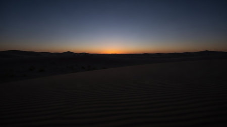 A desert landscape at dusk shows rippled sand dunes with a faint orange sunset on the distant horizon.の素材